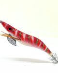 Señuelo jibionera para pesca de cefalópodos, diseño realista con cuerpo segmentado, ojos prominentes y corona de anzuelos múltiples. Ideal para atraer sepias y calamares en aguas costeras. Modelo color rojo con patrón rayas blancas, naranjas y blancas, plumas laterales y aleta metálica para mayor estabilidad.