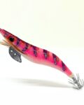 Señuelo jibionera para pesca de cefalópodos, diseño realista con cuerpo segmentado, ojos prominentes y corona de anzuelos múltiples. Ideal para atraer sepias y calamares en aguas costeras. Modelo rosa fluor con patrón rayas lilas y rojas, plumas laterales y aleta metálica para mayor estabilidad.